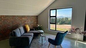 A modern living room with a brick wall, a large window with a view of a field and mountains, a grey sofa, a blue chair, and a glass coffee table. - World of Horses Flats - Lucky (Stellenbosch)
