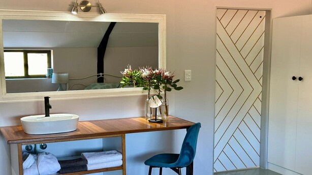 Une salle de bain avec un meuble-lavabo en bois, un lavabo blanc, un miroir, une chaise bleue et une porte ornée d’un motif géométrique.