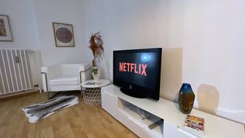 Ein modernes Wohnzimmer mit einem weißen Sofa, einem Fernseher, der 'NETFLIX' anzeigt, einem Heizkörper, einer Vase mit Trockenpflanzen und einer Zeitschrift auf einem Beistelltisch.