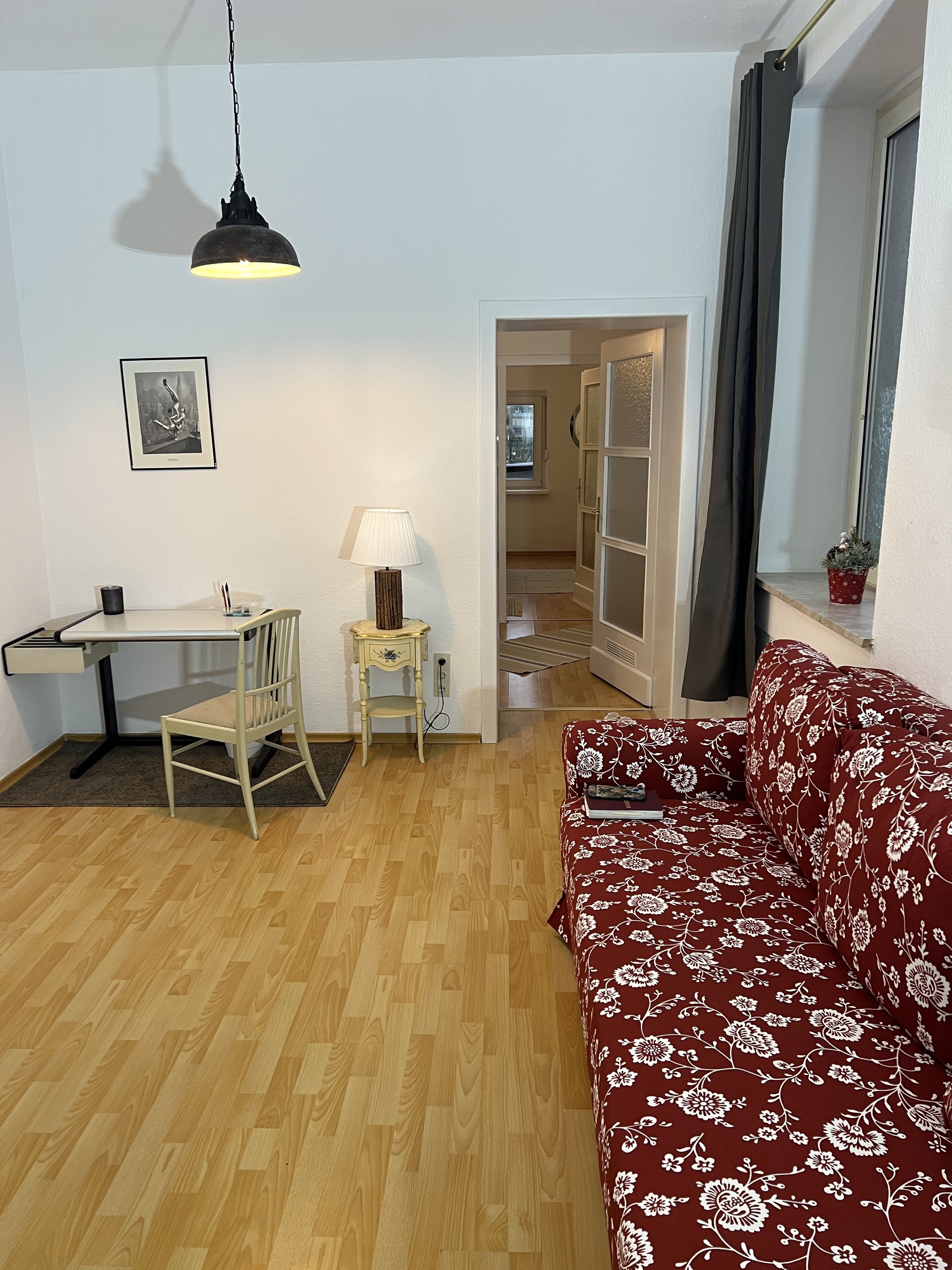 A compact living space with a wooden floor, a desk with a chair, a red patterned sofa, a hanging lamp, and a framed picture on the wall.
