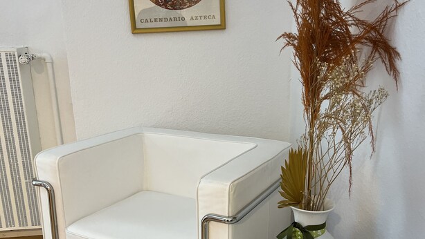 Un sillón blanco de cuero, una mesa redonda blanca con un cuaderno, un jarrón decorativo con plantas secas y un calendario colgado en la pared.
