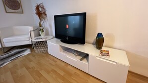 Una sala de estar moderna con un televisor de pantalla plana sobre un mueble blanco, un sofá blanco, una mesita auxiliar con un jarrón y un plato decorativo en la pared.