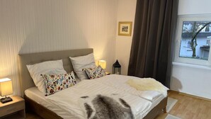 Un dormitorio con una cama grande, dos mesitas de noche, una mesilla auxiliar, una ventana con cortinas y un cuadro en la pared.
