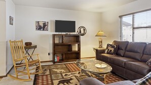 A living room with a brown leather sofa, a glass coffee table, a wooden rocking chair, a bookshelf, a flat-screen TV, and a window with blinds. - Outpost: Vine Street 2 - Close to Snow King! (Jackson)