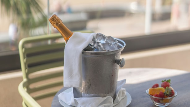 Une table avec une bouteille de champagne dans un seau à glace, un saladier de fruits et une vue sur une plage et l’océan.