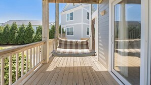 A wooden deck with a swing chair, a glass door, and a house in the background. - Corolla & Lime: Oceanside | Resort Amenities (Corolla)