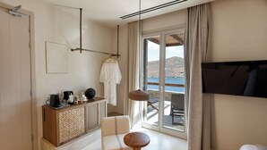 A modern hotel room with a balcony, a TV mounted on the wall, a wooden cabinet with a woven texture, a white sofa, and a round wooden table. - Yfes (Kithnos)