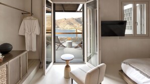 A bedroom with a bed, a chair, a small round table, a robe hanging on a rack, and a TV mounted on the wall. - Yfes (Kithnos)