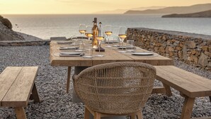 A wooden table set for dining with wine glasses and a bottle, overlooking a rocky beach and the sea. - Yfes (Kithnos)