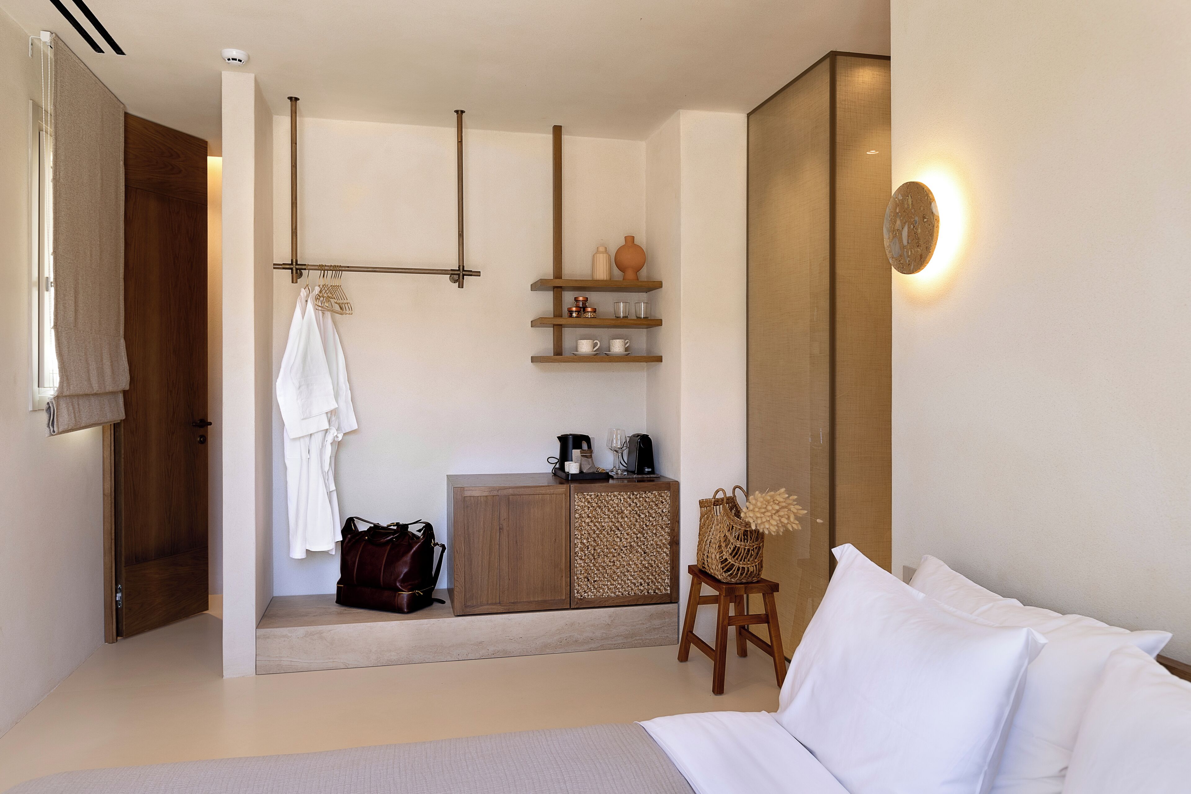 A modern hotel room with a bed, a wooden wardrobe, a small table with a basket, and a shelf with cups and a vase.