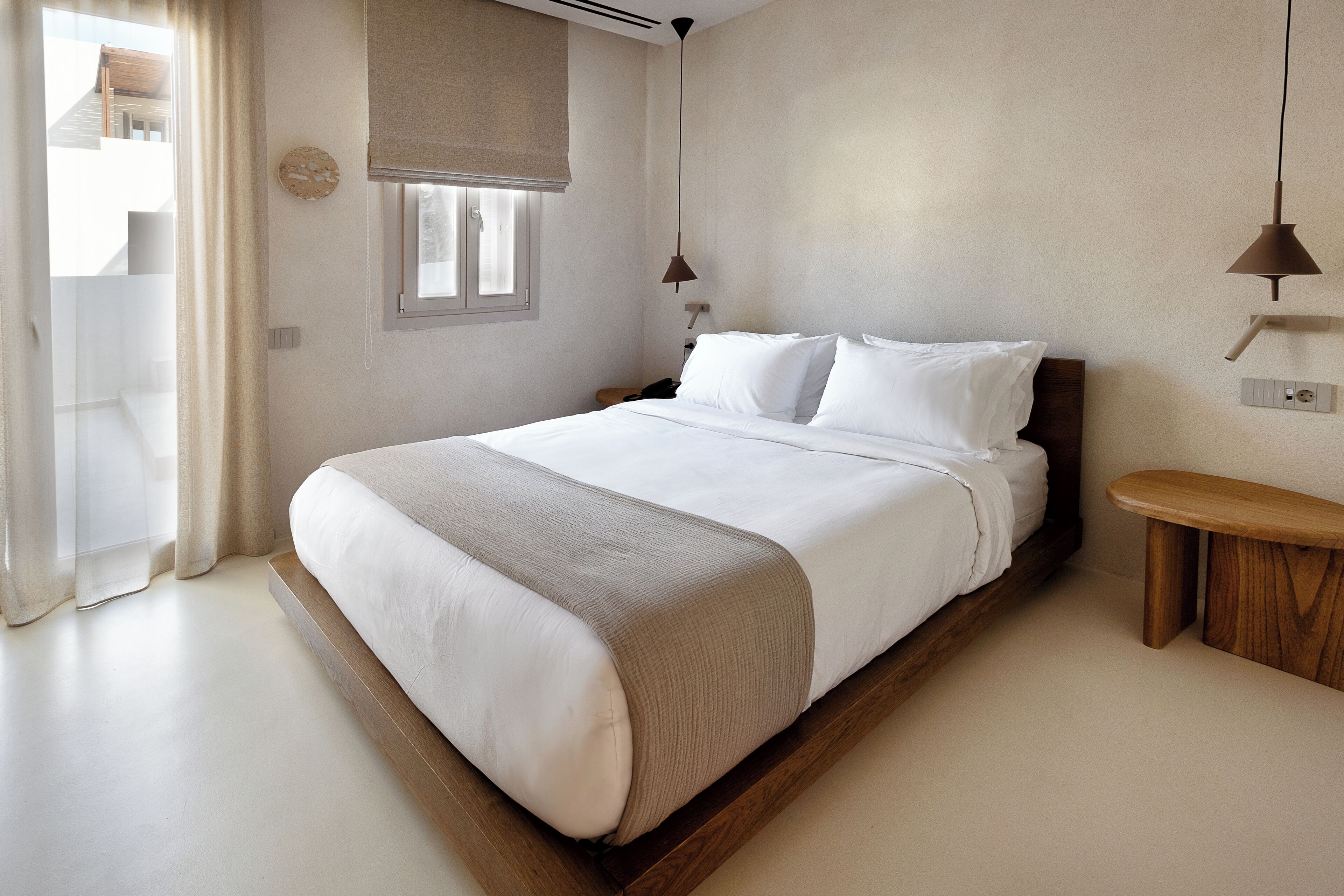 A bedroom with a large bed, a bedside table, a hanging light, and a window with a view of the outside.