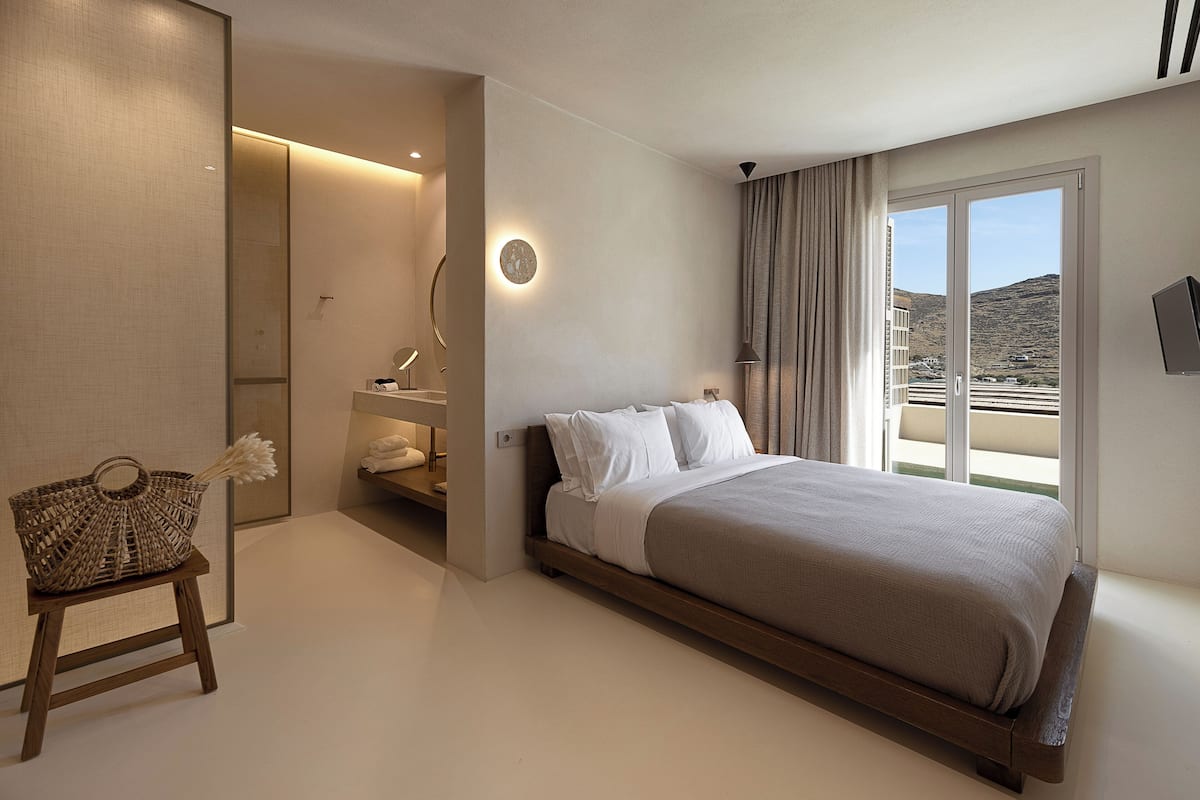A modern bedroom with a large bed, a small seating area, a bedside table, and a view of the outdoors.