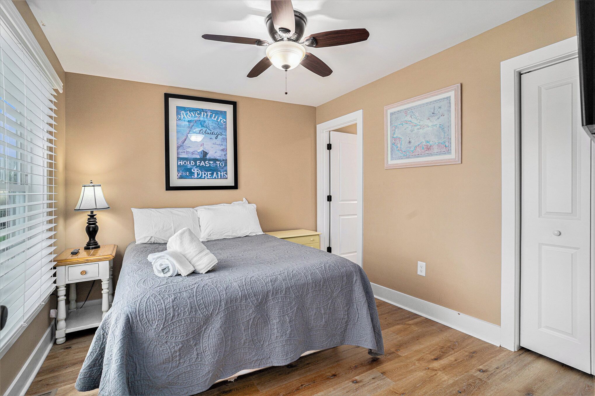 Une chambre à coucher comprenant un lit, un ventilateur de plafond, une table de chevet, une lampe et deux tableaux encadrés accrochés au mur.