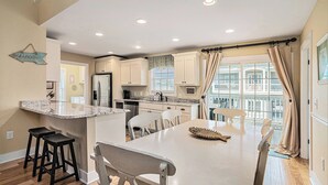 A modern kitchen with white cabinetry, a central island with stools, and a large window with curtains. - On Point: Private Pool Across From Beach (Murrells Inlet)
