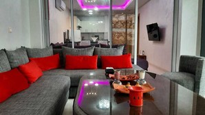 Un salon moderne avec un canapé d’angle gris, des coussins rouges, une table basse en verre agrémentée d’objets décoratifs, et une cuisine en arrière-plan.