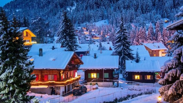 A snow-covered village with wooden houses, some with visible windows and lights, surrounded by snow-laden trees and a mountain in the background.