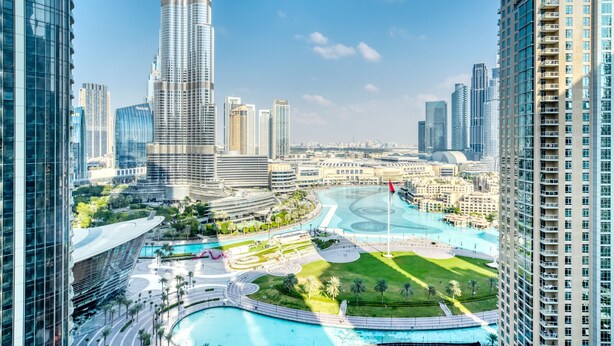 Eine moderne Skyline mit Wolkenkratzern, einem markanten Turm und einer Uferpromenade mit Pool und Grünflächen.