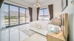 Une chambre moderne avec un grand lit, une table de chevet avec une lampe, une bouteille d’eau et une vue sur les montagnes par de grandes fenêtres.