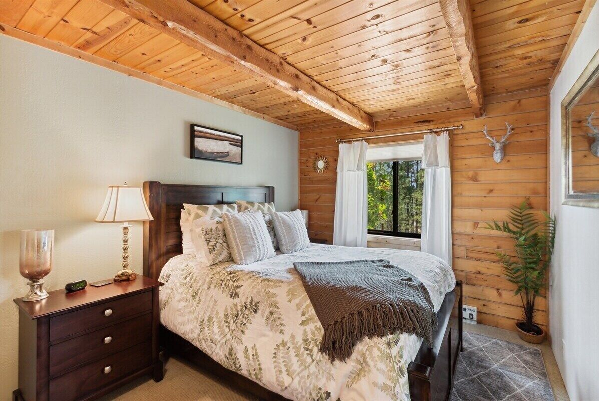 A bedroom with a wooden ceiling, a bed with a floral bedspread, a nightstand with a lamp, and a window with curtains.