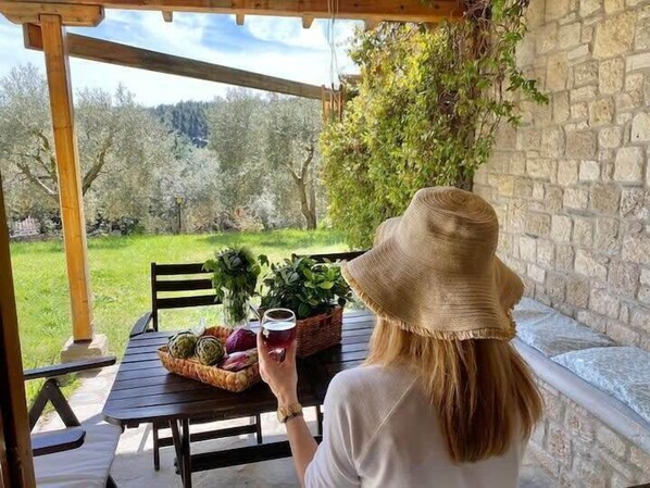 A person seated at a wooden table with a basket of fruits and a glass of red drink, overlooking a stone wall and a grassy area with trees.