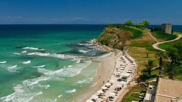 A beach with a row of sun loungers and umbrellas, a clear view of the ocean, and a stone structure on the hillside.