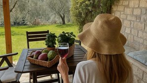 Een vrouw zit aan een buitentafel met een mand vol fruit en een glas rode wijn, met uitzicht op een tuin met stenen muren en begroeiing.