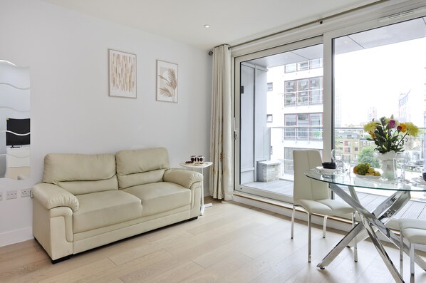 A modern living room with a beige sofa, a glass dining table, and a view of the city through large windows.