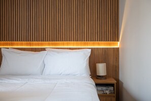 Een opgemaakt bed met witte lakens en kussens, een houten hoofdbord met verticale latten, en een nachtkastje met een lamp en boeken.