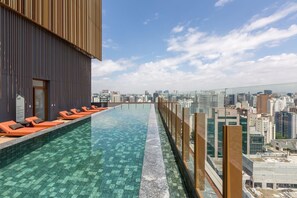Une piscine à débordement située sur le toit, avec des chaises longues orange, une balustrade en verre et une vue panoramique sur la ville.