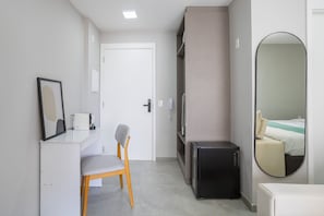 A hotel room with a white desk, a chair, a black cabinet, a mirror, and a bed with a white and green striped bedspread.