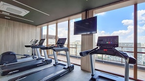 Une salle de sport équipée de tapis de course et d’un vélo d’appartement, de grandes fenêtres offrant une vue sur la ville et d’un téléviseur fixé au mur.