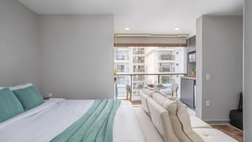 Quarto moderno com cama grande, sofá e varanda com vista para outros edifícios.