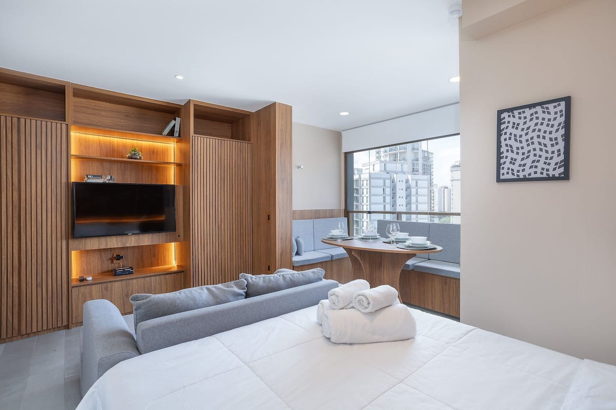 Une chambre d’hôtel moderne dotée d’un grand lit, d’un coin salon avec un canapé et des fauteuils, d’une armoire en bois avec des étagères intégrées, d’une télévision à écran plat et d’un balcon avec une table et des chaises.
