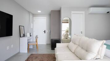 Quarto de hotel moderno com sofá branco, escrivaninha com cadeira, TV de tela plana na parede e um aparelho de ar condicionado pequeno.