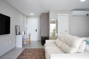 Une chambre d’hôtel moderne avec un canapé blanc, un bureau avec une chaise, un téléviseur à écran plat fixé au mur et un petit climatiseur.