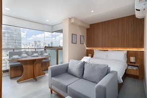 A modern hotel room with a city view, a wooden accent wall, a grey sofa, a wooden dining table, and a bed with white linens. - D'Oru Vila Nova Conceição (São Paulo)