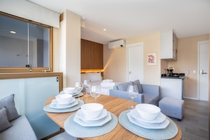 Een moderne, goed verlichte kamer met een eettafel voor vier personen, een bank, een kitchenette en een groot raam.