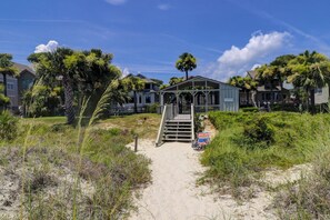 A beach house with a covered porch, a sandy pathway leading to it, and a sign reading "Oceanwood Beach House." - Sunset Rentals | Oceanwood 101 (Hilton Head Island)
