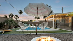 A backyard with a pool, two white chairs, a fire pit, and a large striped umbrella. - Old Town Mid-Century Mod - Heated Pool (Scottsdale)