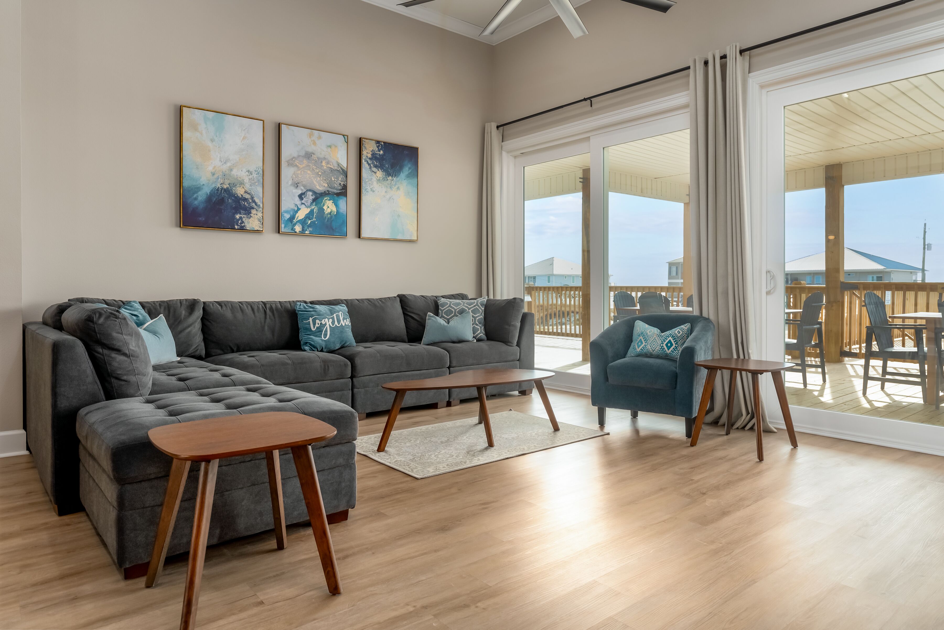 Un salon moderne avec un canapé d’angle gris, une petite table en bois et un fauteuil bleu. Les murs sont ornés de tableaux abstraits et une grande porte-fenêtre coulissante donne accès à une terrasse extérieure.