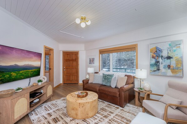 A modern living room with a wooden ceiling, a large flat-screen TV, a brown leather sofa, a round wooden coffee table, and a beige armchair.