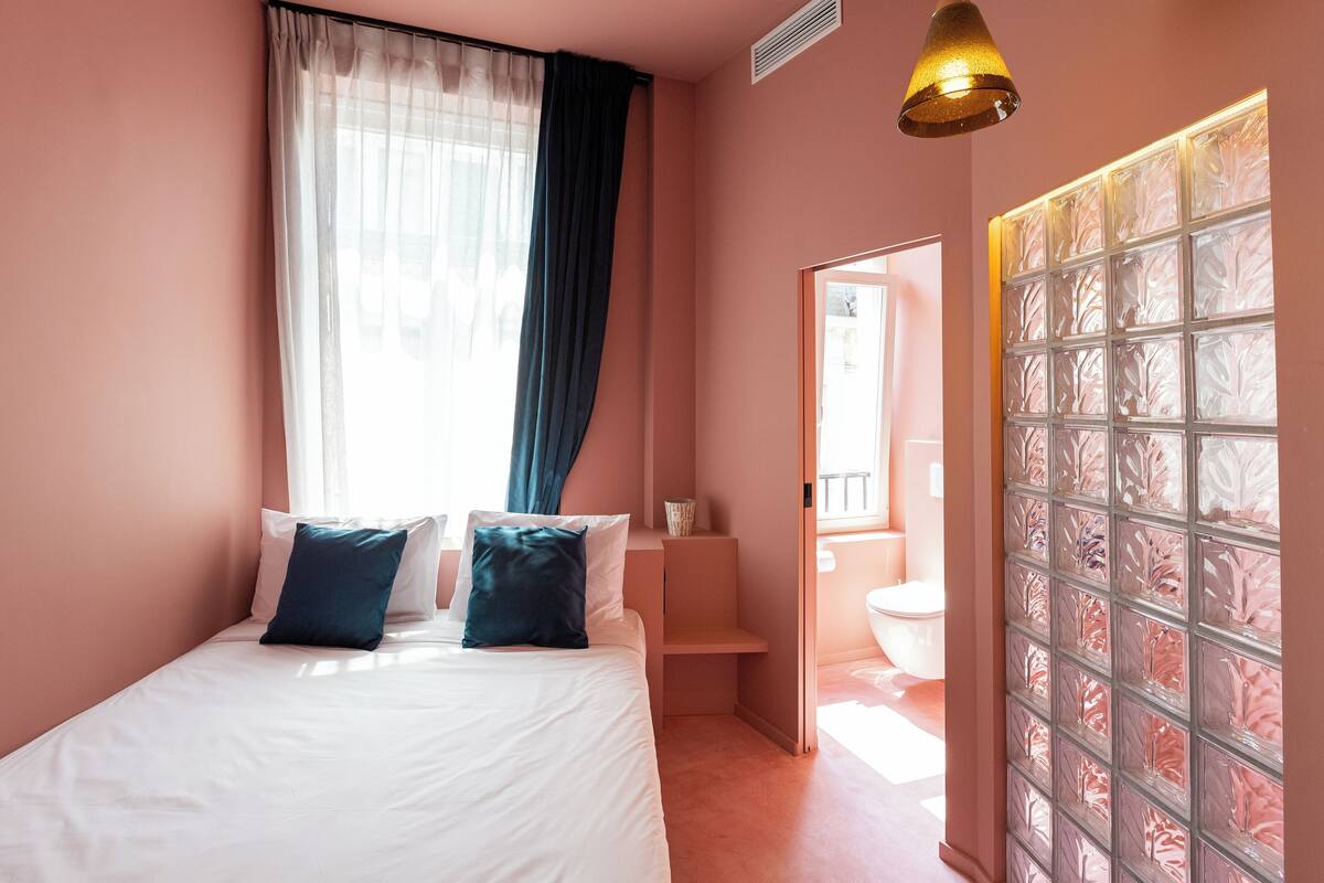 A bedroom with a bed, a window with sheer curtains, a bedside table, and a bathroom door with frosted glass panels.