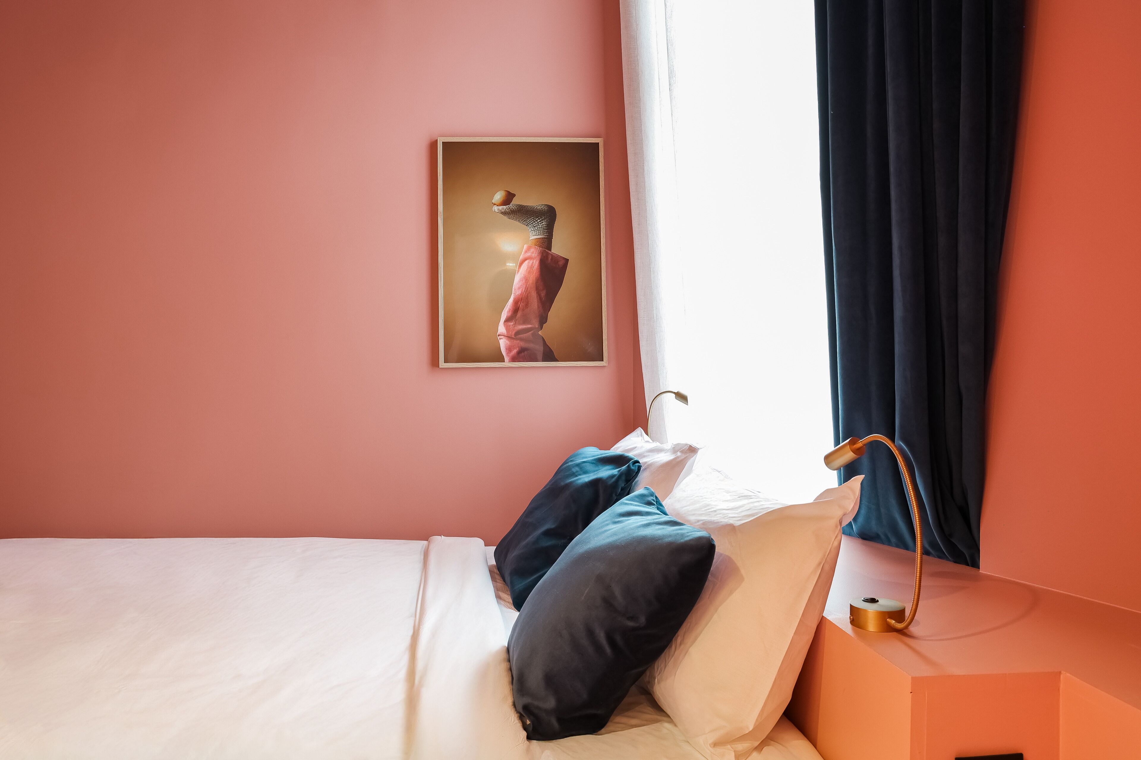 A neatly made bed with white and dark pillows, a framed picture on the wall, and a bedside table with a lamp.