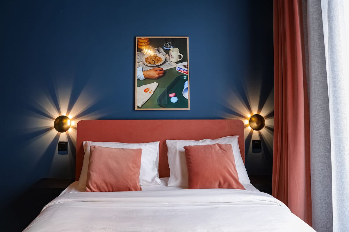 A hotel room with a bed, two pillows, a nightstand, a wall-mounted lamp, and a painting on the wall.