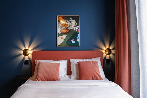 Habitación de hotel con cama, dos almohadas, mesita de noche, lámpara de pared y un cuadro en la pared.