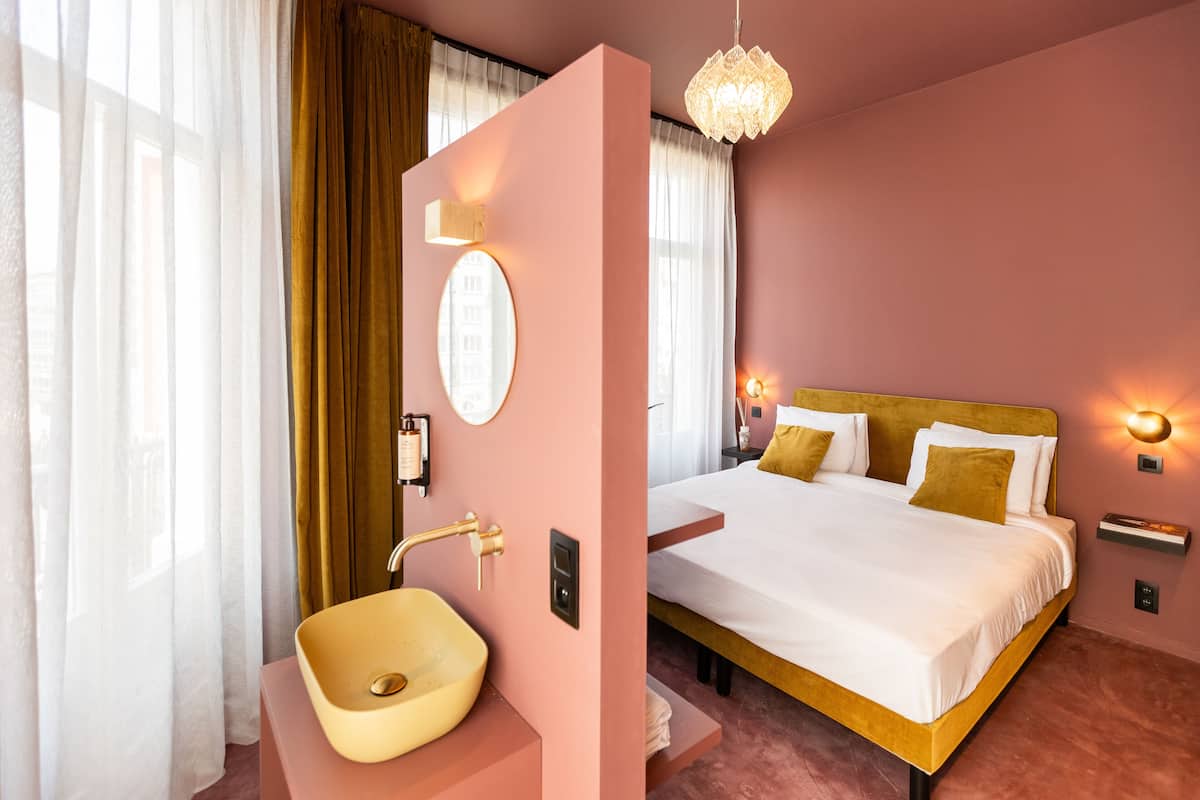 A hotel room with a bed, a bedside table, a wall-mounted light, a mirror, and a sink.