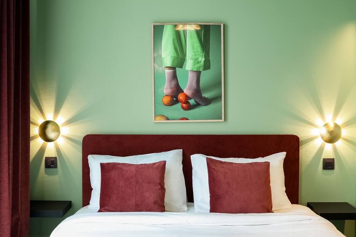 A neatly made bed with a red headboard and white bedding, a framed artwork above it, and two wall-mounted lamps on either side.