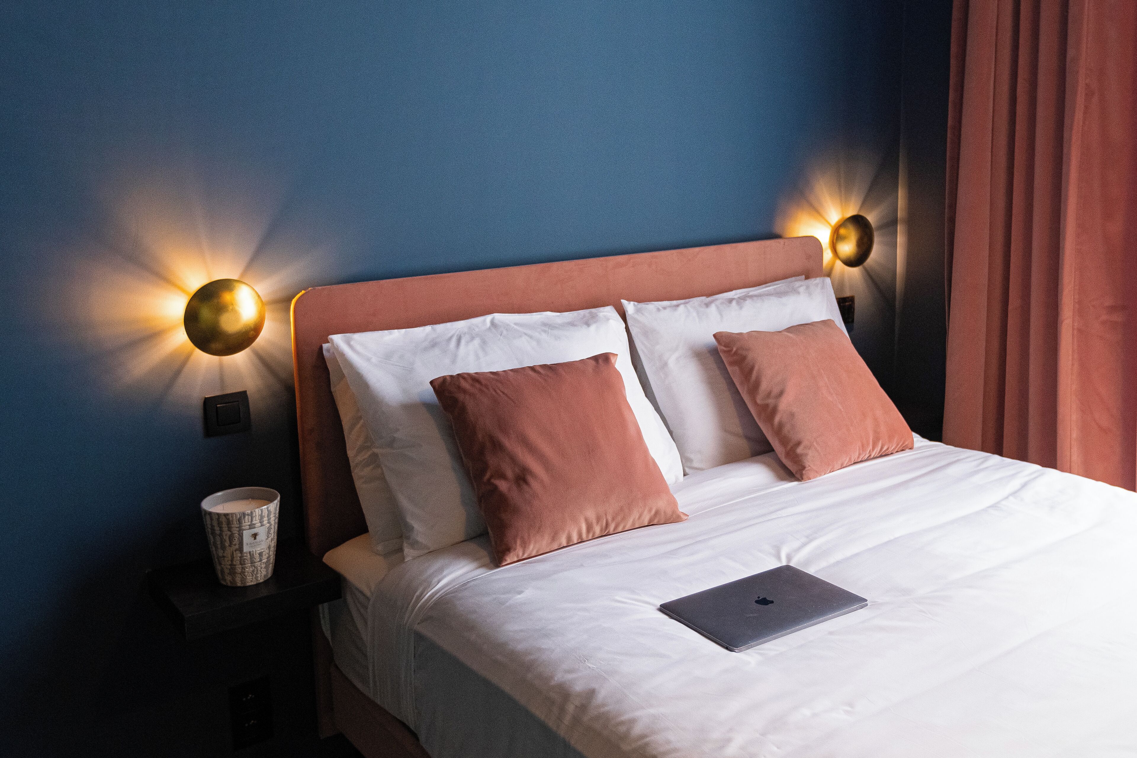 A neatly made bed with white and brown pillows, a laptop on the bed, and two wall-mounted lamps on either side.