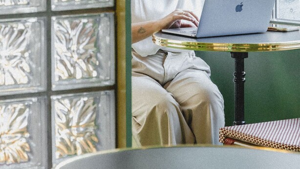 A person using a laptop at a table with a notebook beside it, in a room with a green wall and decorative glass panels. - The Mila Brussels (Brussels)