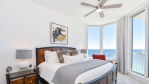 Un dormitorio con una cama grande, un ventilador de techo, una mesita de noche, una lámpara y una ventana con vistas al océano.